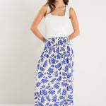 A model wears a long white skirt with a bold blue floral print, elastic waistband, and relaxed fit, paired with a white tank top and neutral sandals.