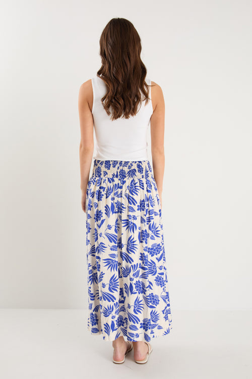 A model wears a long white skirt with a bold blue floral print, elastic waistband, and relaxed fit, paired with a white tank top and neutral sandals.