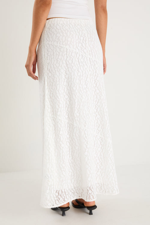 A model wears a long white lace maxi skirt with a plain, lined design, styled with a fitted white tank top and black sandals.