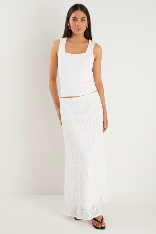 A model wears a long white lace maxi skirt with a plain, lined design, styled with a fitted white tank top and black sandals.