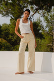 Woman wearing a white tank top and beige pants standing outdoors with greenery in the background