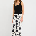 Woman wearing an ivory bias-cut midi skirt with black floral print, elastic waist, and pull-on fit, styled with black sandals and a tucked-in black top.