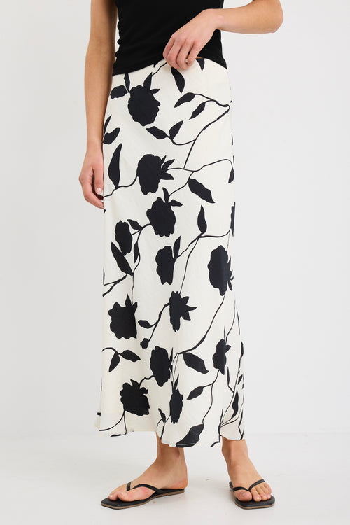 Woman wearing an ivory bias-cut midi skirt with black floral print, elastic waist, and pull-on fit, styled with black sandals and a tucked-in black top.