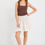 Woman wearing a natural tiered linen-blend mini skirt with a shirred waistband and layered hem, styled with a chocolate tank.