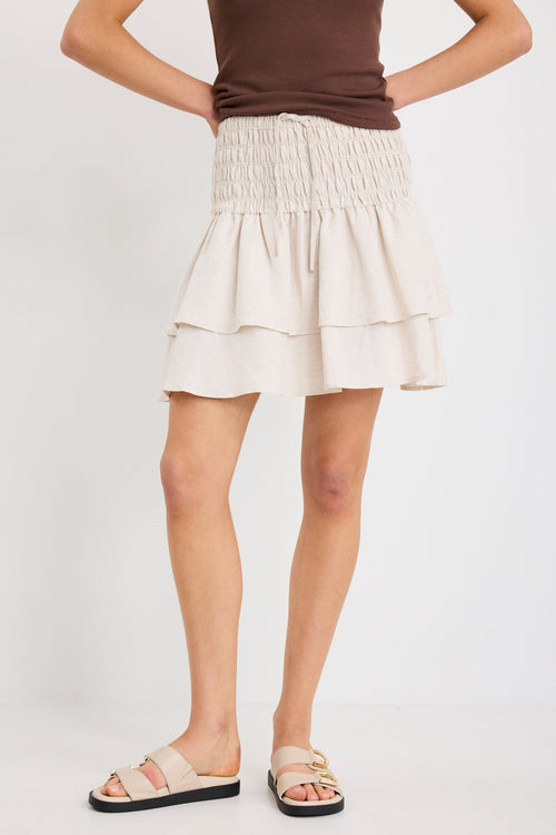 Woman wearing a natural tiered linen-blend mini skirt with a shirred waistband and layered hem, styled with a chocolate tank.