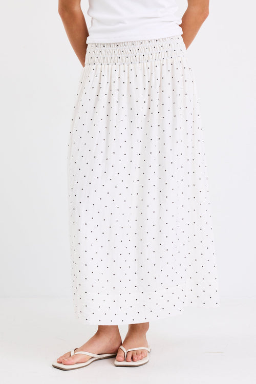 Woman wearing a cream maxi skirt with black polka dots and a shirred elastic waistband, paired with a white tank top and sandals, standing against a white background.