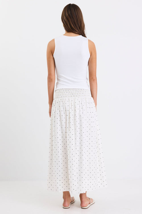 Woman wearing a cream maxi skirt with black polka dots and a shirred elastic waistband, paired with a white tank top and sandals, standing against a white background.