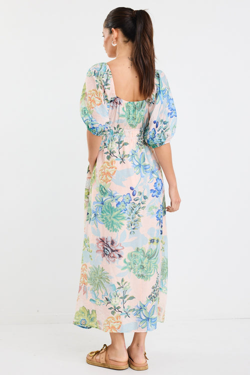 woman wearing a flowing floral puff sleeve dress with elastic waist and pastel blue and green print, styled with tan sandals.