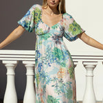 Woman wearing a floaty floral maxi dress in pastel blues, pinks and greens with puff sleeves and a gathered waist.