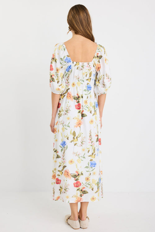 Woman wearing a cream midi dress with a sweetheart neckline, puff sleeves, elastic waist, and garden floral print in red, yellow, and blue tones.