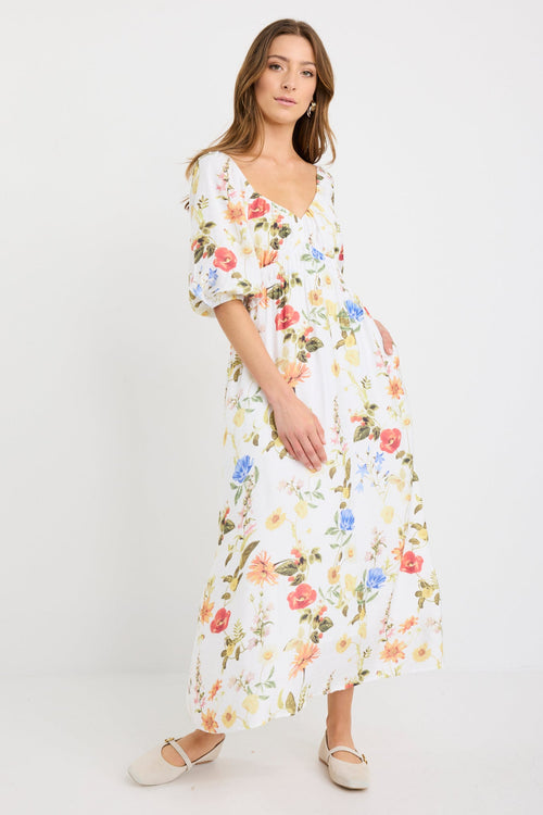 Woman wearing a cream midi dress with a sweetheart neckline, puff sleeves, elastic waist, and garden floral print in red, yellow, and blue tones.