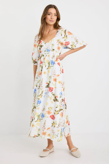 Woman wearing a cream midi dress with a sweetheart neckline, puff sleeves, elastic waist, and garden floral print in red, yellow, and blue tones.