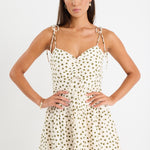 Woman wearing a cream mini dress with khaki polka dots, tiered skirt and tie-up straps, styled with gold jewellery.