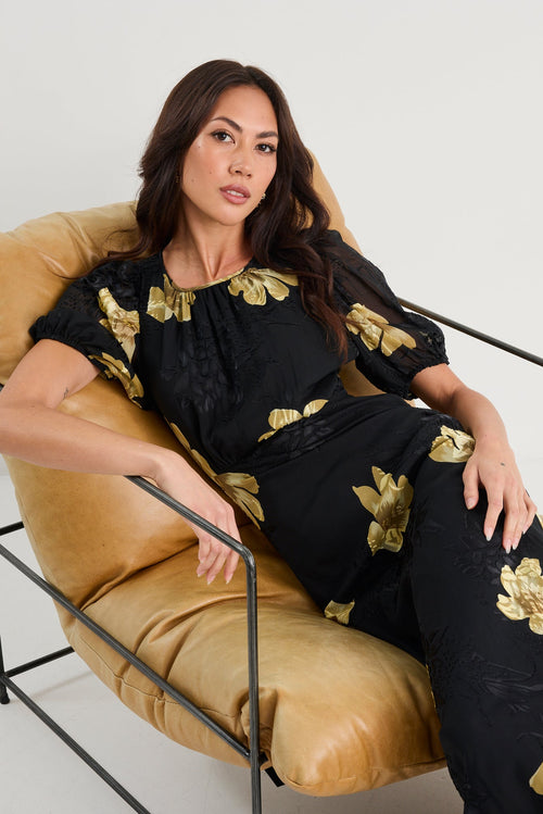 Woman sitting on a chair wearing a black outfit with gold floral patterns.