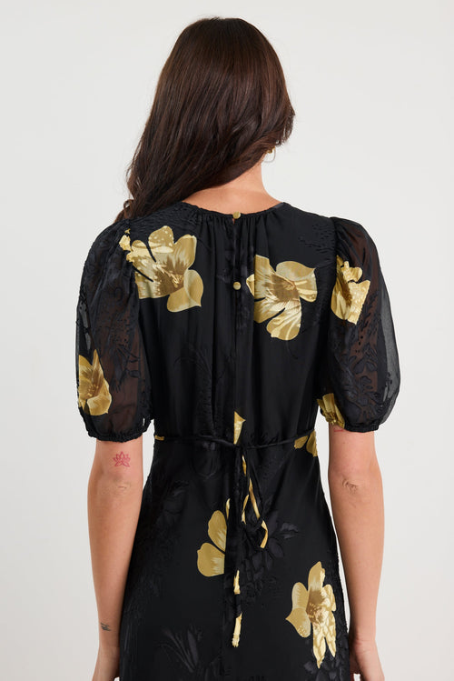 Woman wearing a black dress with gold floral patterns from behind on a white background