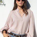 A woman wearing a light pink and white vertically striped long-sleeve blouse with a scalloped V-neckline, tucked into light-wash blue jeans with a brown belt, posing against a white stucco wall.
