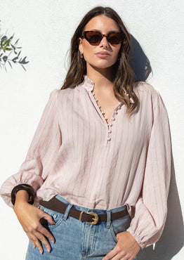 A woman wearing a light pink and white vertically striped long-sleeve blouse with a scalloped V-neckline, tucked into light-wash blue jeans with a brown belt, posing against a white stucco wall.
