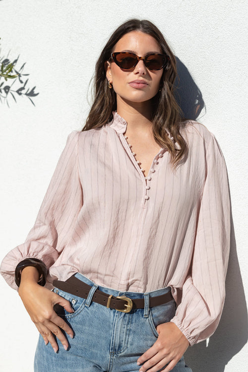 A woman wearing a light pink and white vertically striped long-sleeve blouse with a scalloped V-neckline, tucked into light-wash blue jeans with a brown belt, posing against a white stucco wall.