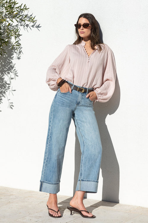 A woman stands against a white wall wearing a light pink long-sleeve blouse, light blue wide-leg jeans, a black belt, and black sandals.