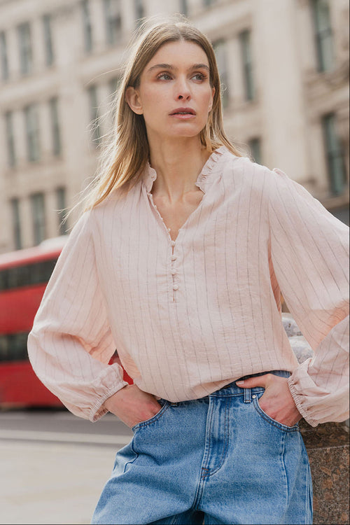 Woman wearing a light pink blouse and blue jeans standing in an urban setting with a red bus in the background.