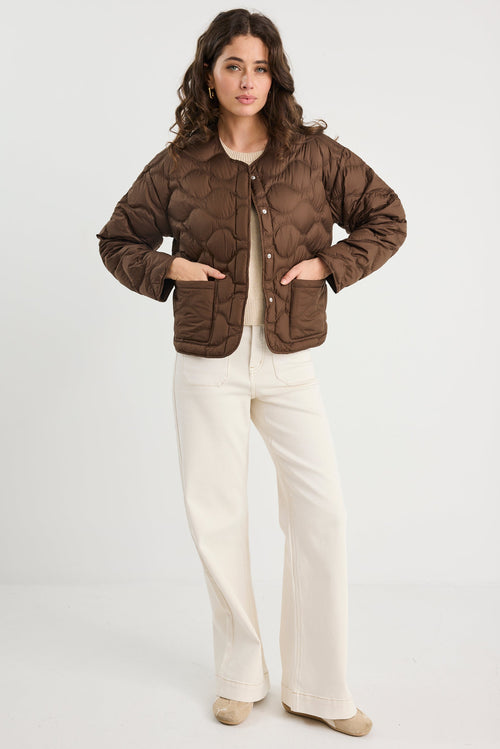 Woman wearing a brown quilted jacket and white pants on a white background