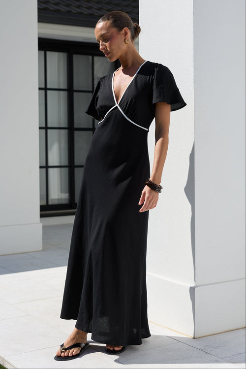 A woman wearing a black maxi dress with white contrast piping stands outdoors in front of a modern white building with columns.