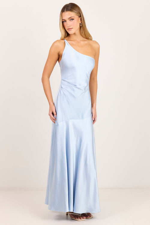 A woman modeling a light blue one-shoulder maxi dress with a draped bodice and flowing skirt, standing against a plain off-white studio background.