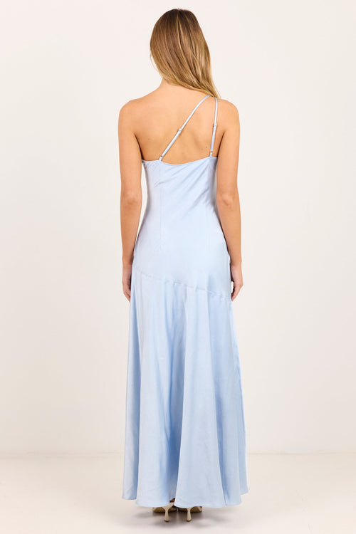A woman viewed from the back wearing a light blue one-shoulder maxi dress with thin straps and a gathered skirt, standing against a plain off-white studio background.
