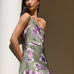 A woman wearing a one-shoulder olive green dress with a purple floral print, posing next to a white column in warm, natural light.