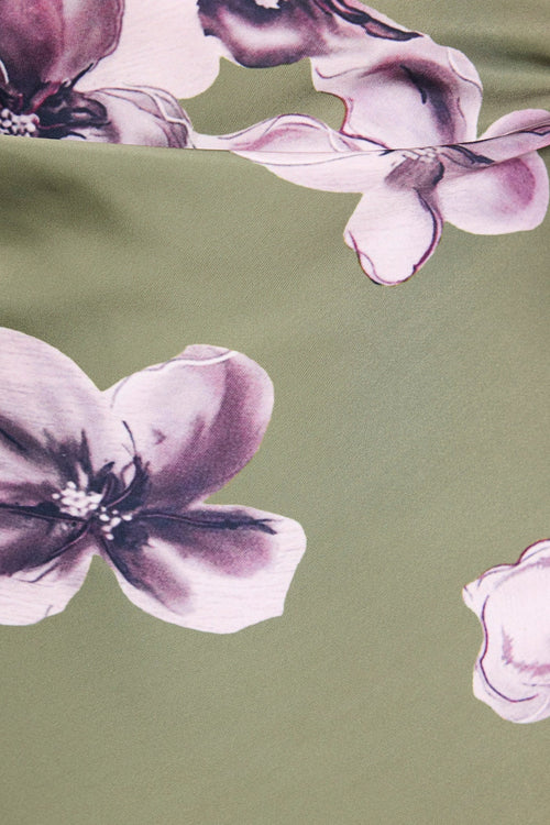 A close-up of a fabric surface with a large floral print. The background is a muted sage green, decorated with oversized watercolor-style flowers in lavender, mauve, and pale pink tones.