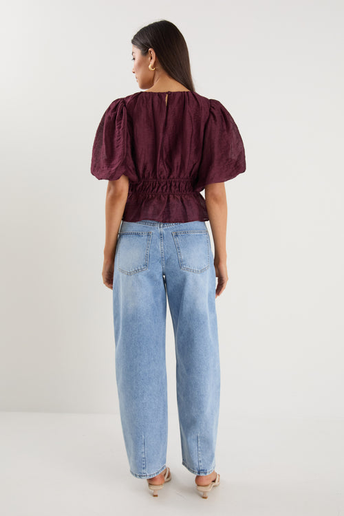 A model wears a burgundy puff sleeve top with a gathered waist, styled with light blue high-waisted jeans.