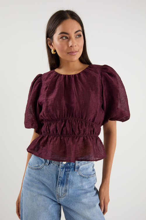 A model wears a burgundy puff sleeve top with a gathered waist, styled with light blue high-waisted jeans.