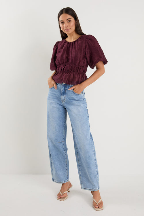 A model wears a burgundy puff sleeve top with a gathered waist, styled with light blue high-waisted jeans.