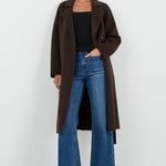 Woman wearing a brown coat, black top, and blue jeans on a white background