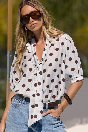 A woman wearing a white short-sleeve button-up blouse with dark brown polka dots, styled with light-wash blue denim shorts, a brown belt, and dark sunglasses.