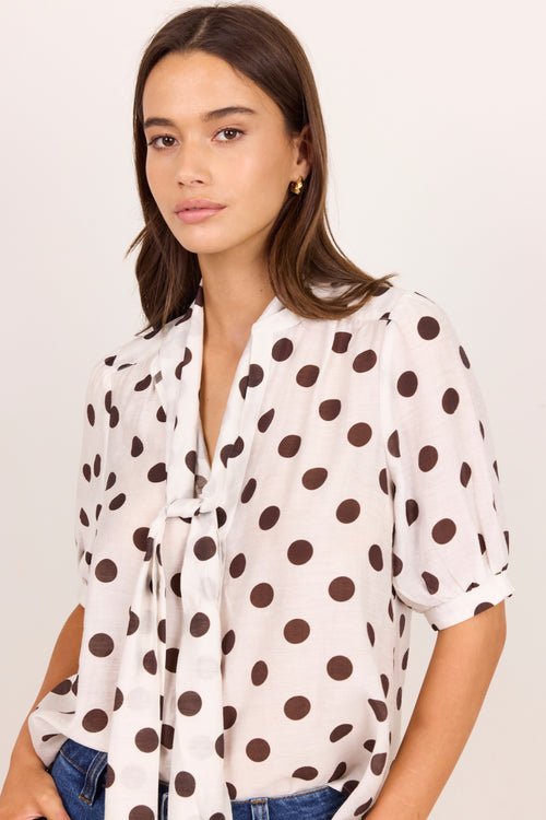 A woman wearing a white short-sleeve blouse with dark brown polka dots and a tie-neck detail, paired with blue jeans, against a plain off-white background.