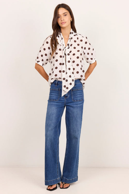 A woman modeling a white short-sleeve button-up shirt with brown polka dots, paired with high-waisted blue flare jeans and black sandals, standing against a plain off-white studio background.