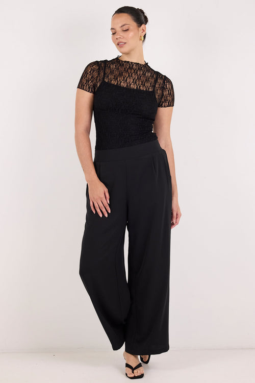 A woman modeling a black short-sleeve jumpsuit with a sheer lace yoke and wide-leg pants, standing against a plain white studio background.