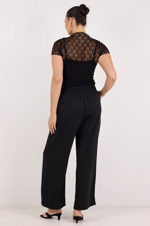 A woman viewed from the back wearing a black short-sleeve jumpsuit with a sheer geometric pattern across the upper back and shoulders, paired with black heeled sandals, against a plain white studio background.