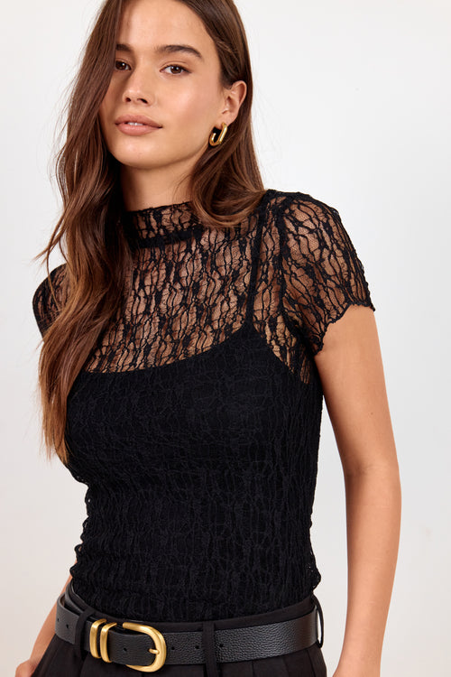 A woman wearing a black lace mock-neck top with short sleeves, paired with a black belt, against a plain light background.