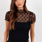 A woman modeling a black short-sleeve top with a sheer floral lace yoke and a textured black knit body, paired with black trousers and a black belt with a gold buckle, against a plain light gray background.