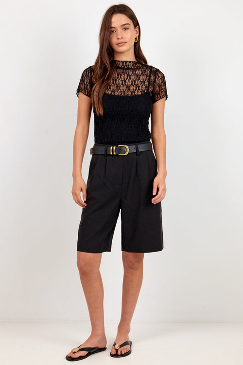 A woman modeling a black short-sleeve top with a sheer geometric pattern and black pleated shorts, styled with a black belt and black sandals, against a plain white studio background.