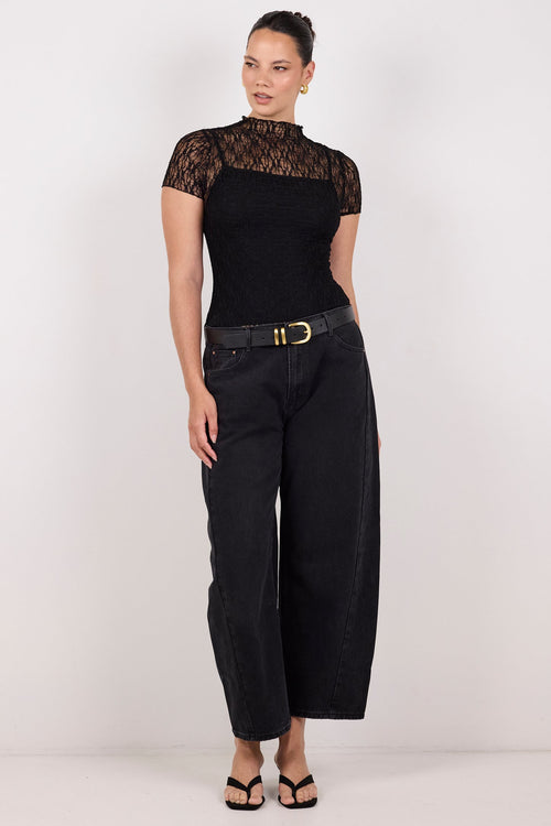 A woman modeling a black short-sleeve top with sheer black lace sleeves and black wide-leg cropped pants with a black belt, standing against a plain light gray studio background.