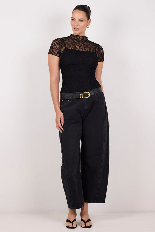 A woman modeling a black short-sleeve top with sheer black lace sleeves and black wide-leg cropped pants with a black belt, standing against a plain light gray studio background.