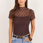 A woman modeling a short-sleeve brown lace and textured knit top with a mock neck, paired with light blue jeans and a brown belt, against a plain off-white background.