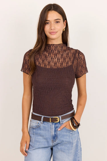 A woman modeling a short-sleeve brown lace and textured knit top with a mock neck, paired with light blue jeans and a brown belt, against a plain off-white background.