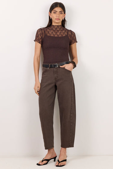 A woman stands against a plain white studio wall wearing a dark brown short-sleeve top with a sheer lace yoke, dark brown wide-leg cropped pants, a black belt, and black heeled sandals.