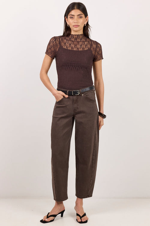 A woman modeling a dark brown short-sleeve top with a sheer lace yoke and matching brown cropped pants, styled with a black belt and black sandals against a plain white studio background.