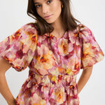 Woman wearing a pink multi floral short sleeve top with bubble sleeves, cinched waist, and crew neckline, styled with blue jeans.