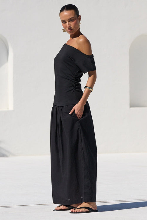 A woman wearing a black off-the-shoulder maxi dress stands on a white tiled poolside patio with a modern white stucco building in the background.